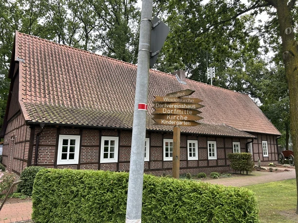 Heimathaus Lahn ©Emsland Tourismus GmbH (1).JPEG Backsteinfachwerkhaus mit rotem Ziegeldach, umgeben von Bäumen und Wegweisern zu Ortszielen.