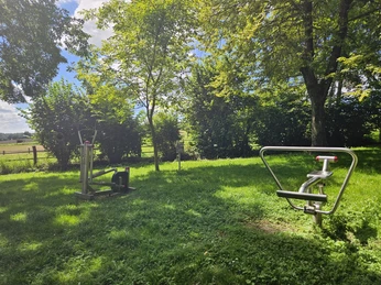Fitnessgeräte auf einer grünen Wiese umgeben von Bäumen unter blauem Himmel im Freien.