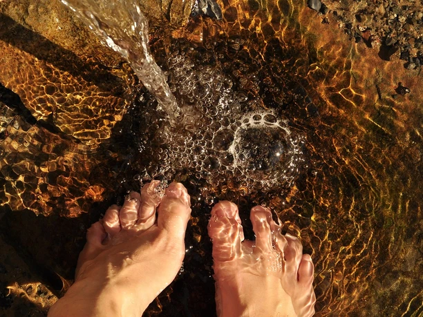Füße in klarem, flachem Wasser, leicht bewegt von einem sanft fließenden Wasserstrahl.
