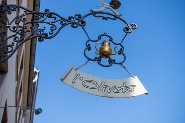 L'Oliveto Schild Schild "l'Oliveto" an verschnörkelter Aufhängung vor blauem Himmel