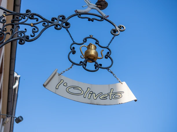 L'Oliveto Schild Schild "l'Oliveto" an verschnörkelter Aufhängung vor blauem Himmel