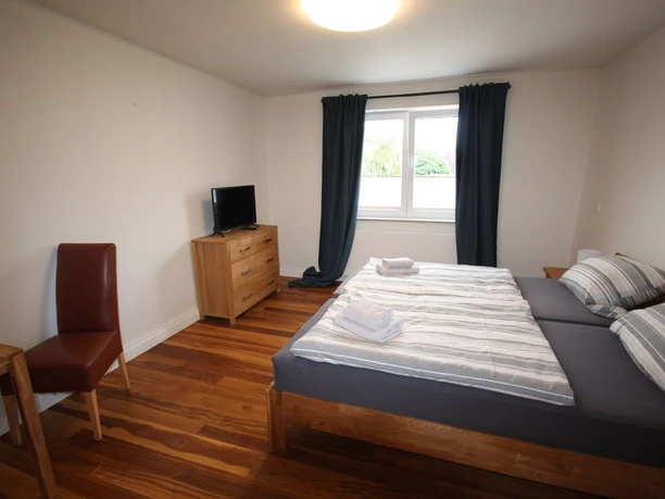 Ein helles, schlicht eingerichtetes Zimmer mit Doppelbett, Holzboden und einem Fenster mit blickdichten Vorhängen.