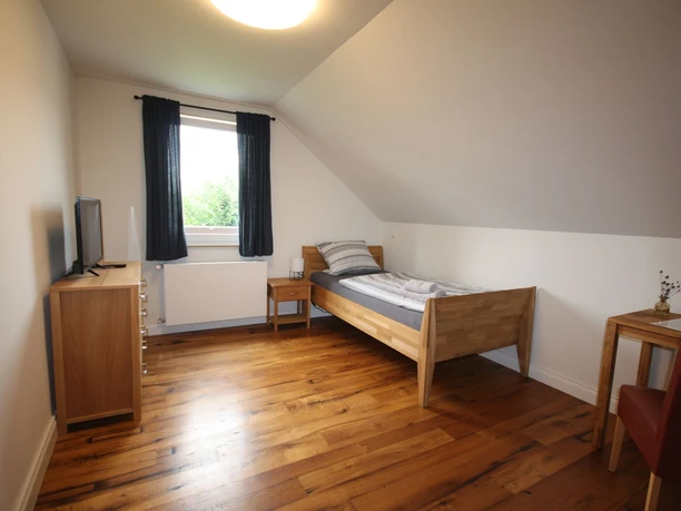 Ein schlichtes Einzelzimmer mit Holzfußboden, Bett, Fernseher, Schreibtisch und einem Fenster.