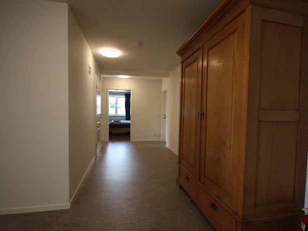 pension_luci_flur1a.jpg Breiter, lichtdurchfluteter Flur mit Holzschrank auf der rechten Seite und Zugang zu einem fensterhellen Raum.