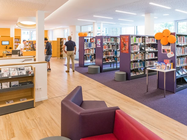 Bibliothek Neuschaefer-Rube.jpg Moderne Bibliothek mit hell erleuchtetem Innenraum; Bücherregale, Sitzgelegenheiten und Lesebereich.