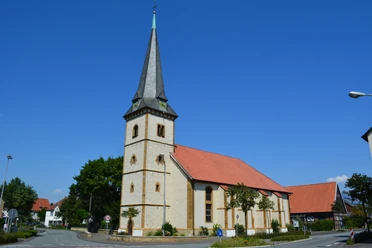Brockhagen - St. Georgskirche Evangelische Kirche mit spitzem Turm und roten Dachziegeln vor blauem Himmel in ländlicher Umgebung.
