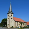 Brockhagen - St. Georgskirche Evangelische Kirche mit spitzem Turm und roten Dachziegeln vor blauem Himmel in ländlicher Umgebung.