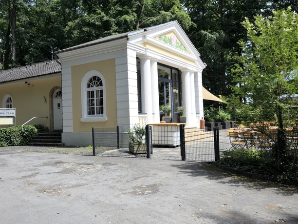 Ein charmantes Gebäude im klassizistischen Stil mit weißen Säulen, vor einem grünen Wald gelegen.