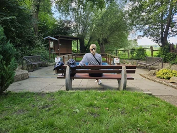 Eine Person auf einer Holzbank, umgeben von grüner Natur, nutzt ein Wassertretbecken im Freien.