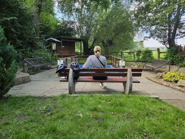 Eine Person auf einer Holzbank, umgeben von grüner Natur, nutzt ein Wassertretbecken im Freien.