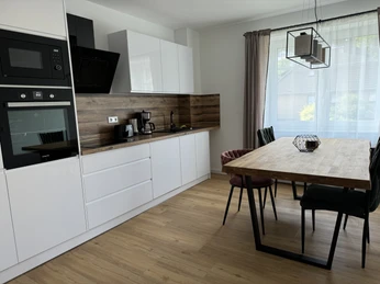 IMG_6380.jpg Moderne, helle Küche mit Esstisch und vier Stühlen, ausgestattet mit Backofen, Kaffeemaschine und viel Tageslicht.