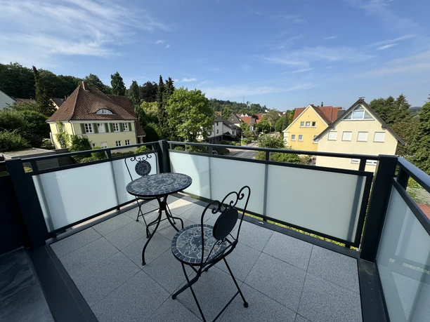 IMG_6447.jpg Zwei schmiedeeiserne Stühle und ein Tisch auf einem Balkon mit Blick auf ein Wohngebiet und Hügel.