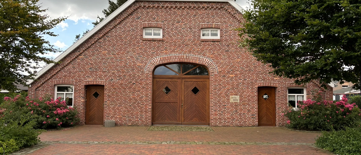 Backsteinbau mit roten Dachziegeln und hölzernen Türen, umgeben von blühenden Büschen und Bäumen.