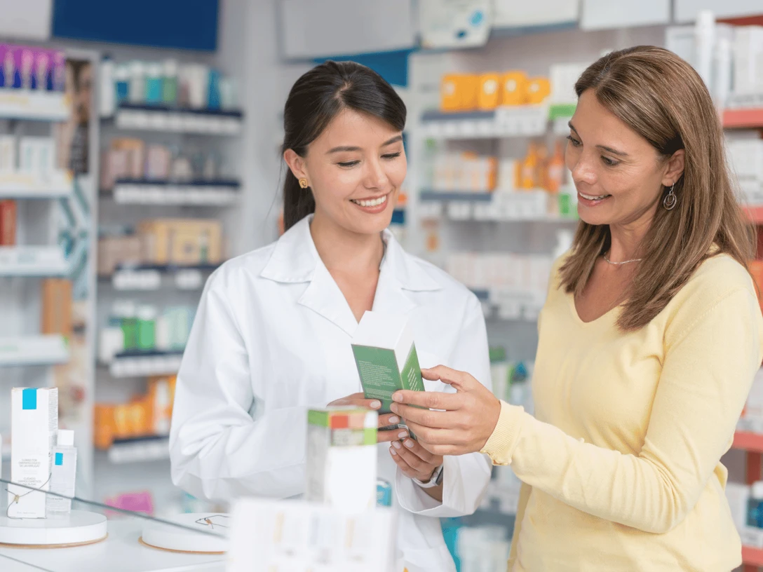 Apotheke Alpha.png Auf dem Bild ist eine Apothekerin zu sehen, die eine Kundin berät. Beide lachen, und die Kundin hält bereits ein Produkt in der Hand. Im Hintergrund ist die Apotheke mit zahlreichen Regalen, die eine Vielzahl von weiteren Produkten zeigen.