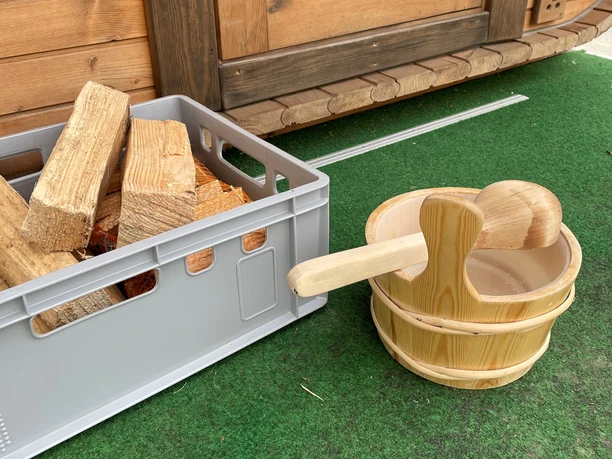 Holzfasseimer mit Schöpfkelle neben Kiste voller Holzscheite, vor hölzerner Fasssauna auf grünem Untergrund.