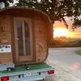 Fasssauna auf Anhänger Fasssauna auf Anhänger vor Sonnenuntergang, umgeben von Bäumen und einer ländlichen Straße.