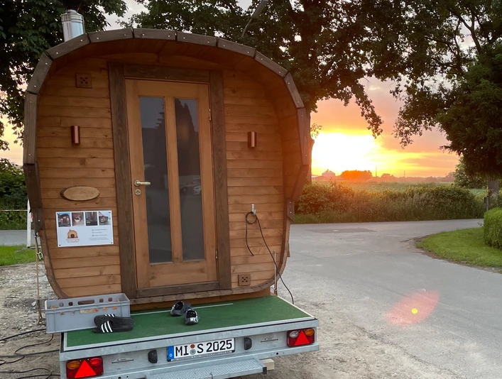 Fasssauna auf Anhänger Fasssauna auf Anhänger vor Sonnenuntergang, umgeben von Bäumen und einer ländlichen Straße.
