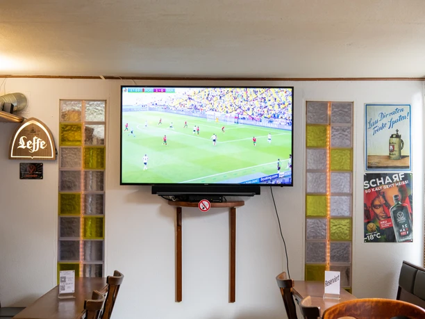 Schloss-Schänke: Innenansicht Ein TV auf dem Fußball läuft hängt an der Wand. Davor stehen Tische und Stühle, an der Wand sind Werbetafeln für Bier und Jägermeister.