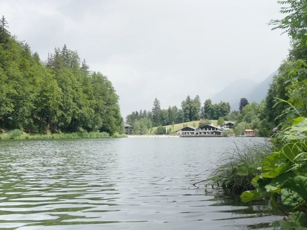 Riessersee See mit grünen Ufern, im Hintergrund ein Gebäude. Wald und Berge bilden die Szenerie.