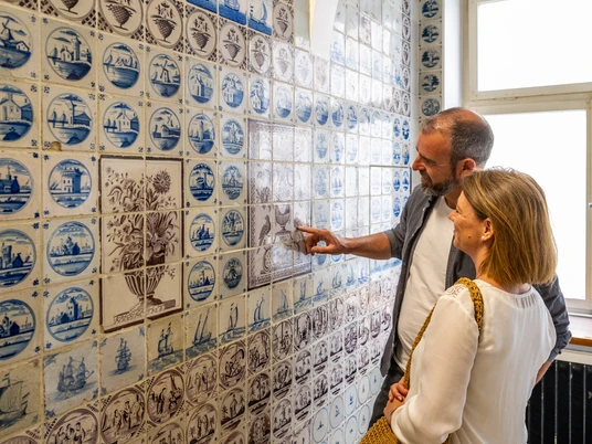 Bielefeld-Delfter-Kacheln-Teutoburger-Wald-Bielefeld-MarketingD-Ketz Mann und Frau betrachten blau-weiße Fliesen mit historischen Motiven in einem lichtdurchfluteten Raum.