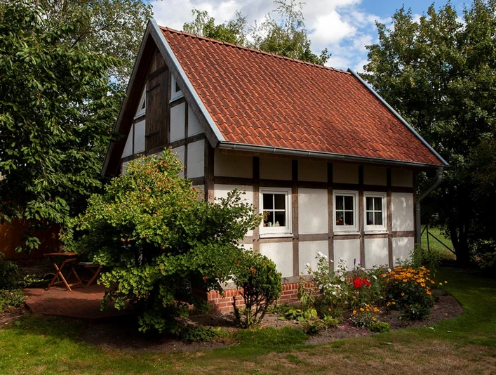 Kleines Fachwerkhaus mit rotem Ziegeldach, umgeben von Bäumen, Sträuchern und einem Blumengarten.