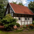 Rahe_Tiny House.jpg Kleines Fachwerkhaus mit rotem Ziegeldach, umgeben von Bäumen, Sträuchern und einem Blumengarten.