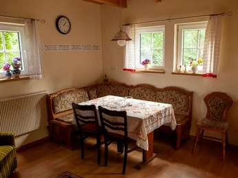 Rahe_Esszimmer.jpg Ein Esszimmer mit Holzmöbeln, bestickter Tischdecke, zwei Fenstern mit Blumenschmuck und Uhr an der Wand.