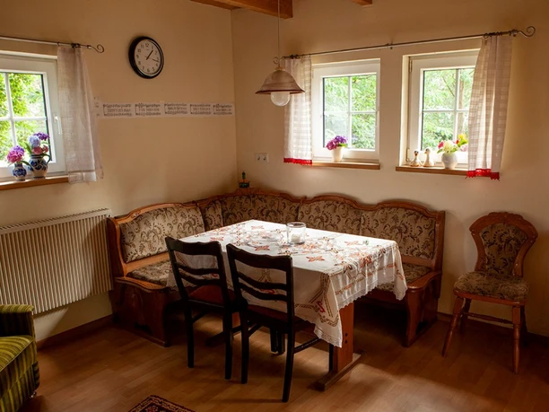 Rahe_Esszimmer.jpg Ein Esszimmer mit Holzmöbeln, bestickter Tischdecke, zwei Fenstern mit Blumenschmuck und Uhr an der Wand.