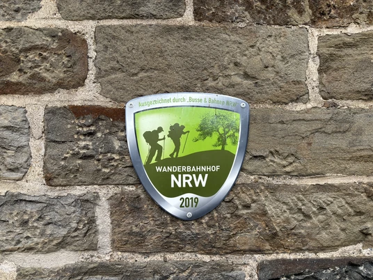 Auszeichnung Wanderbahnhof NRW Schild an einer Steinmauer mit der Aufschrift "Wanderbahnhof NRW 2019", grün und silbern gestaltet.
