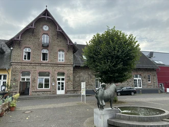 Kultur- und Wanderbahnhof Altes Backsteingebäude mit Uhrturm, Baum und Bronzeskulptur vor einem Wasserspielplatz bei bewölktem Himmel.
