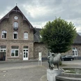 Kultur- und Wanderbahnhof Altes Backsteingebäude mit Uhrturm, Baum und Bronzeskulptur vor einem Wasserspielplatz bei bewölktem Himmel.