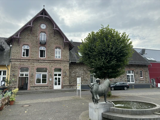 Kultur- und Wanderbahnhof Altes Backsteingebäude mit Uhrturm, Baum und Bronzeskulptur vor einem Wasserspielplatz bei bewölktem Himmel.