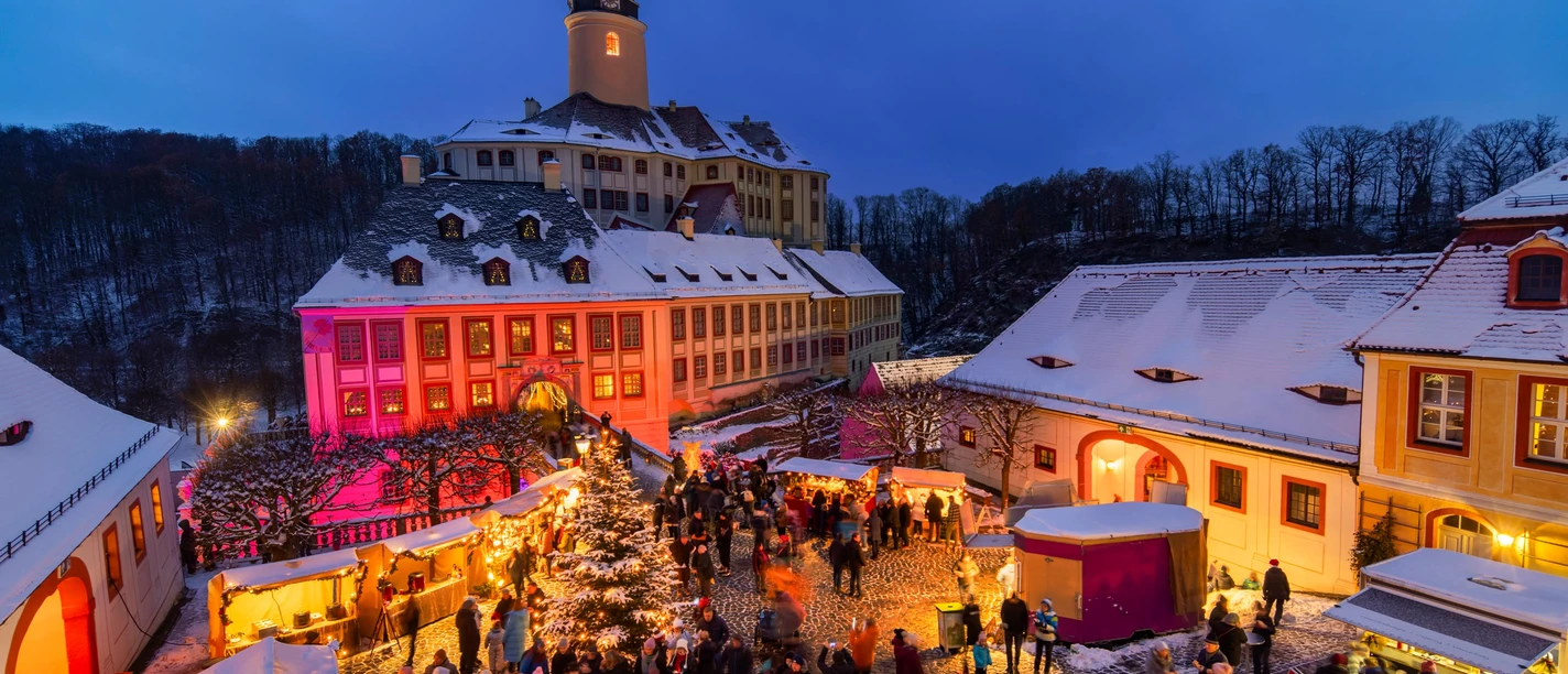 Weesensteiner Schlossweihnacht Belebter Weihnachtsmarkt vor dem beleuchteten Schloss Weesenstein, umgeben von verschneiten Gebäuden.