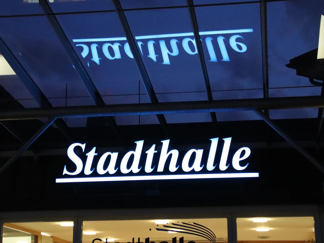 Delbrueck_Stadthalle_Leuchtzug ©Stadthalle Delbrück.JPG Beleuchtetes Stadthalle-Schild bei Nacht mit reflektierendem Glasdach im Vordergrund.