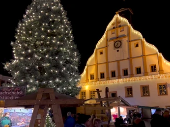 Ein Fest der Sinne - Grimmaer Weihnachtsmarkt Zu sehen ist das Grimmaer Rathaus weihnachtlich beleuchtet. Im Vordergrund steht ein großer geschmückter Weihnachtsbaum und Stände mit Spezialitäten.