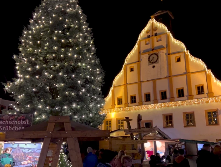 Ein Fest der Sinne - Grimmaer Weihnachtsmarkt Zu sehen ist das Grimmaer Rathaus weihnachtlich beleuchtet. Im Vordergrund steht ein großer geschmückter Weihnachtsbaum und Stände mit Spezialitäten.