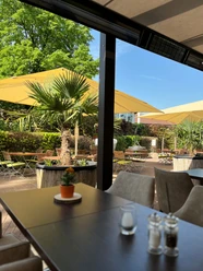 Oase Biergarten.jpg Sonnige Gartenterrasse mit gelben Sonnenschirmen, Palmen und Holztischen in entspannter Atmosphäre.