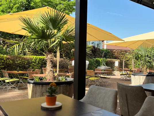 Oase Biergarten.jpg Sonnige Gartenterrasse mit gelben Sonnenschirmen, Palmen und Holztischen in entspannter Atmosphäre.