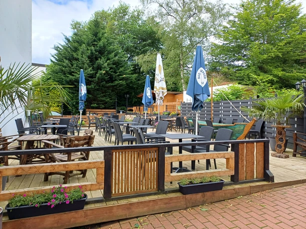 Sonnige Außenterrasse mit Holzmöbeln, Sonnenschirmen und blühenden Pflanzen, umgeben von hohen Bäumen.