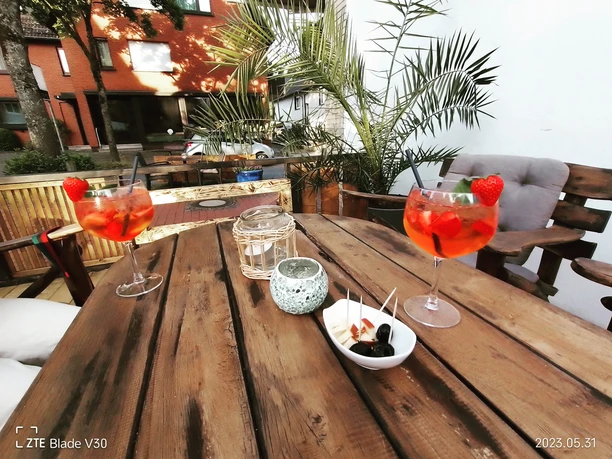 IMG_20230531_200206_690_©Eiscafé Mona Lisa.jpg Zwei Aperol Spritz mit Erdbeerdekor auf Holztisch, umgeben von Palmen und Holzstühlen im sonnigen Freien.