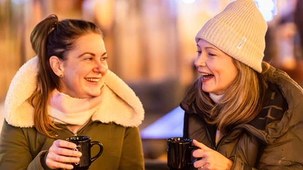 Zusehen sind zwei Frauen die jeweils eine Glühweintasse in der Hand halten und lachen.