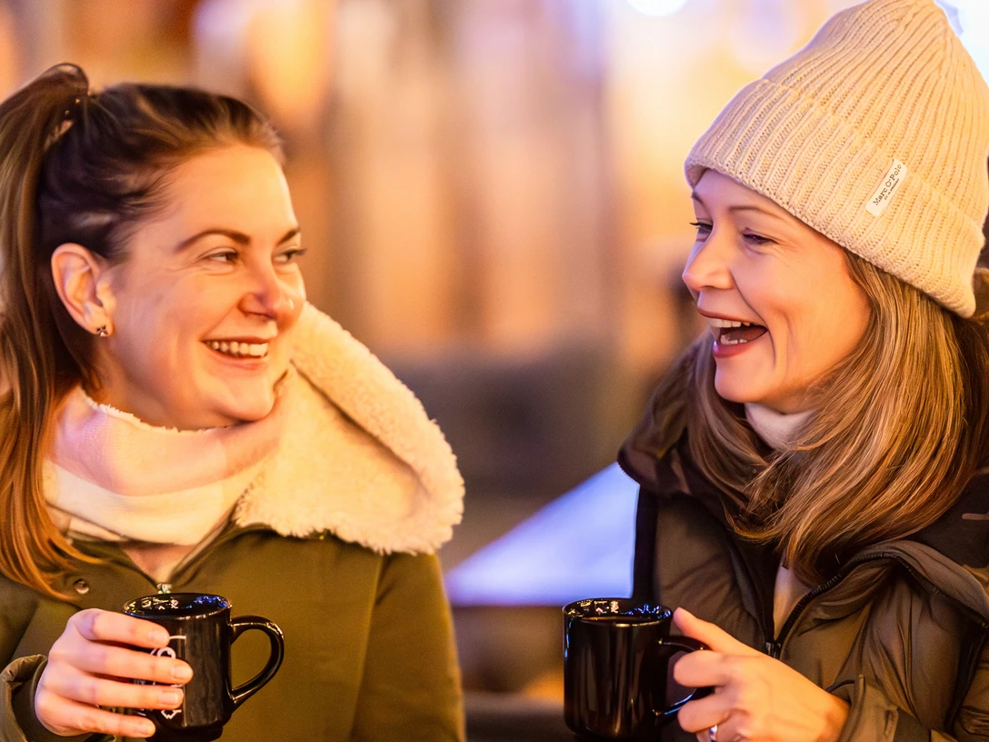 Glühwein trinken auf dem Weihnachtsmarkt Zusehen sind zwei Frauen die jeweils eine Glühweintasse in der Hand halten und lachen.
