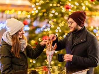Wintermarkt bei Herr Kauzig - Weihnachten in Leipzig Im Vordergrund essen zwei Personen in winterlicher Kleidung Churros und trinken Glühwein auf dem kauzigen Wintermarkt während im Hintergrund Tannenbaum festlich erleuchtet.
