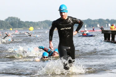 Ein Triathlet in schwarzem Neoprenanzug läuft dynamisch aus dem Wasser, während andere im Hintergrund weiterschwimmen. Die Sonne scheint, und die Wettkampfenergie ist spürbar.