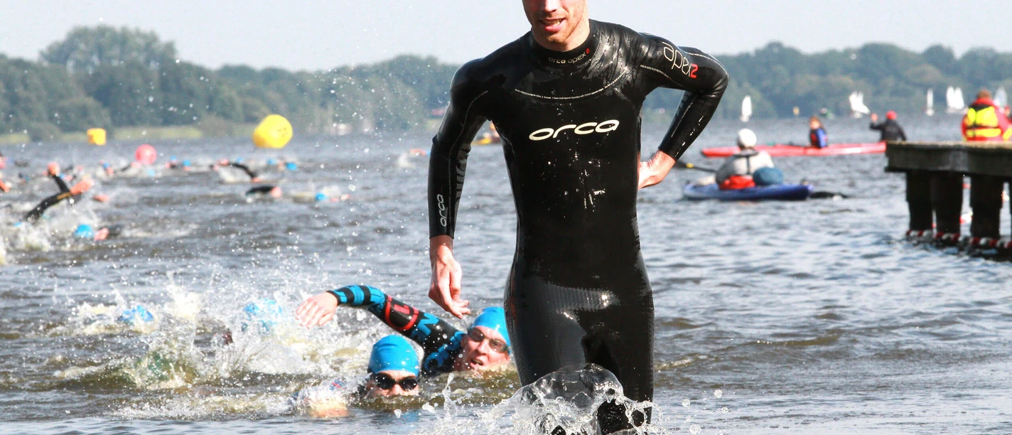 Ein Triathlet in schwarzem Neoprenanzug läuft dynamisch aus dem Wasser, während andere im Hintergrund weiterschwimmen. Die Sonne scheint, und die Wettkampfenergie ist spürbar.