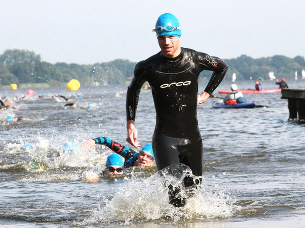 Ein Triathlet in schwarzem Neoprenanzug läuft dynamisch aus dem Wasser, während andere im Hintergrund weiterschwimmen. Die Sonne scheint, und die Wettkampfenergie ist spürbar.