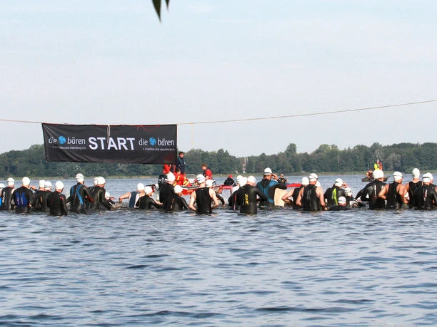Baeren5178.JPG Teilnehmer eines Triathlons stehen im Wasser am Start unter einem Banner, umgeben von einem See in einer natürlichen Umgebung.