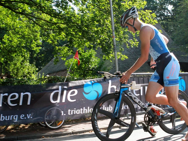 baerentriathlon-radfahren Ein sportlicher Triathlet, im blauen Rennanzug und Helm, schiebt sein Fahrrad bei einem sonnigen Event mit grüner Kulisse.