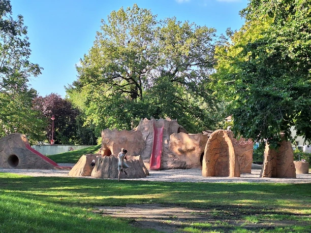 12 - Supervulkan-Zentrum R├Âcknitz - Spielplatz Fred Porphyrstein - Foto Kathleen Schramm.jpg Abenteuerspielplatz mit Kletterstrukturen aus Steinimitat unter schattigen Bäumen.