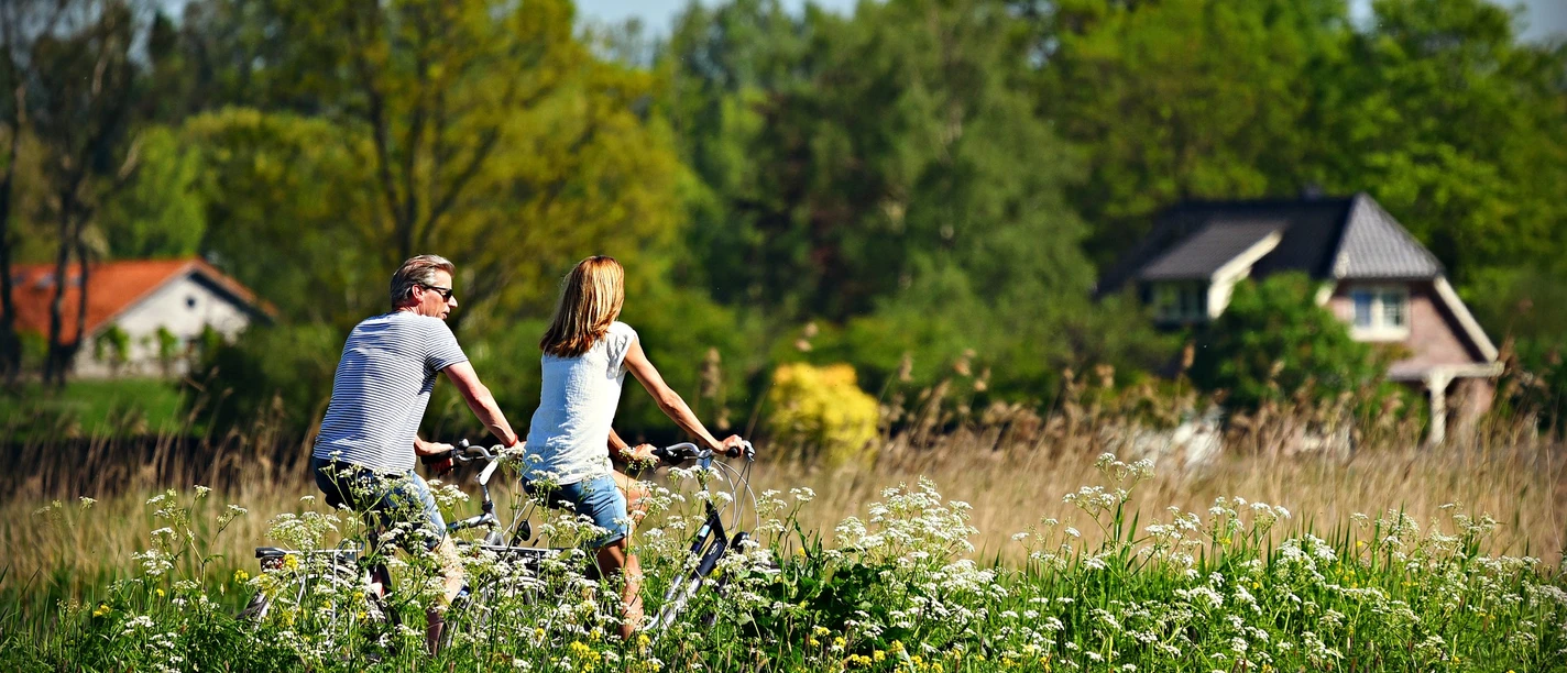radfahren Zwei Personen radeln durch eine blühende Wiese mit Häusern und grünen Bäumen im Hintergrund.
